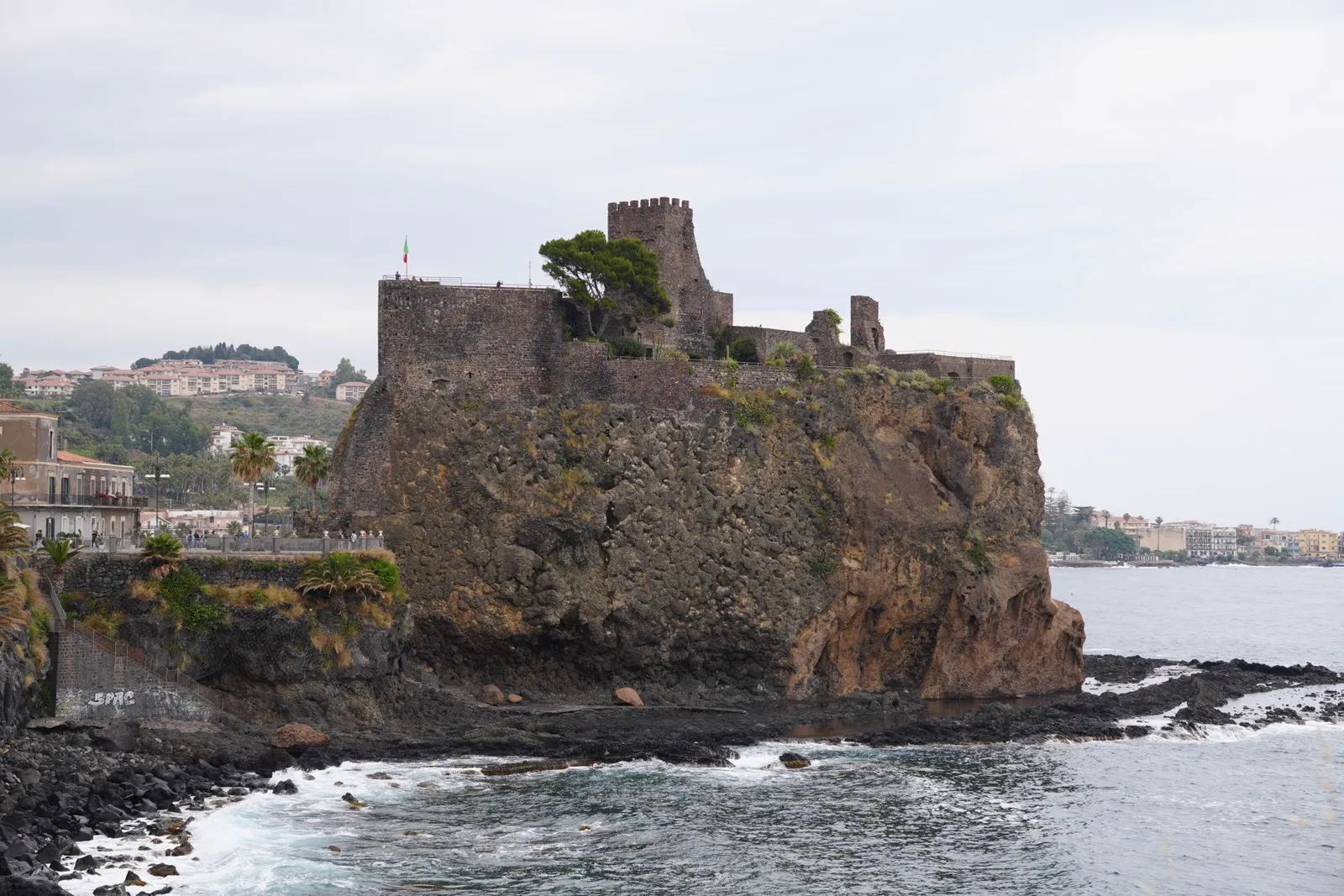 The black lava Norman castle of Aci Castello perched on a rocky basalt promontory over the sea near Catania, Sicily, with the coastline stretching behind it.