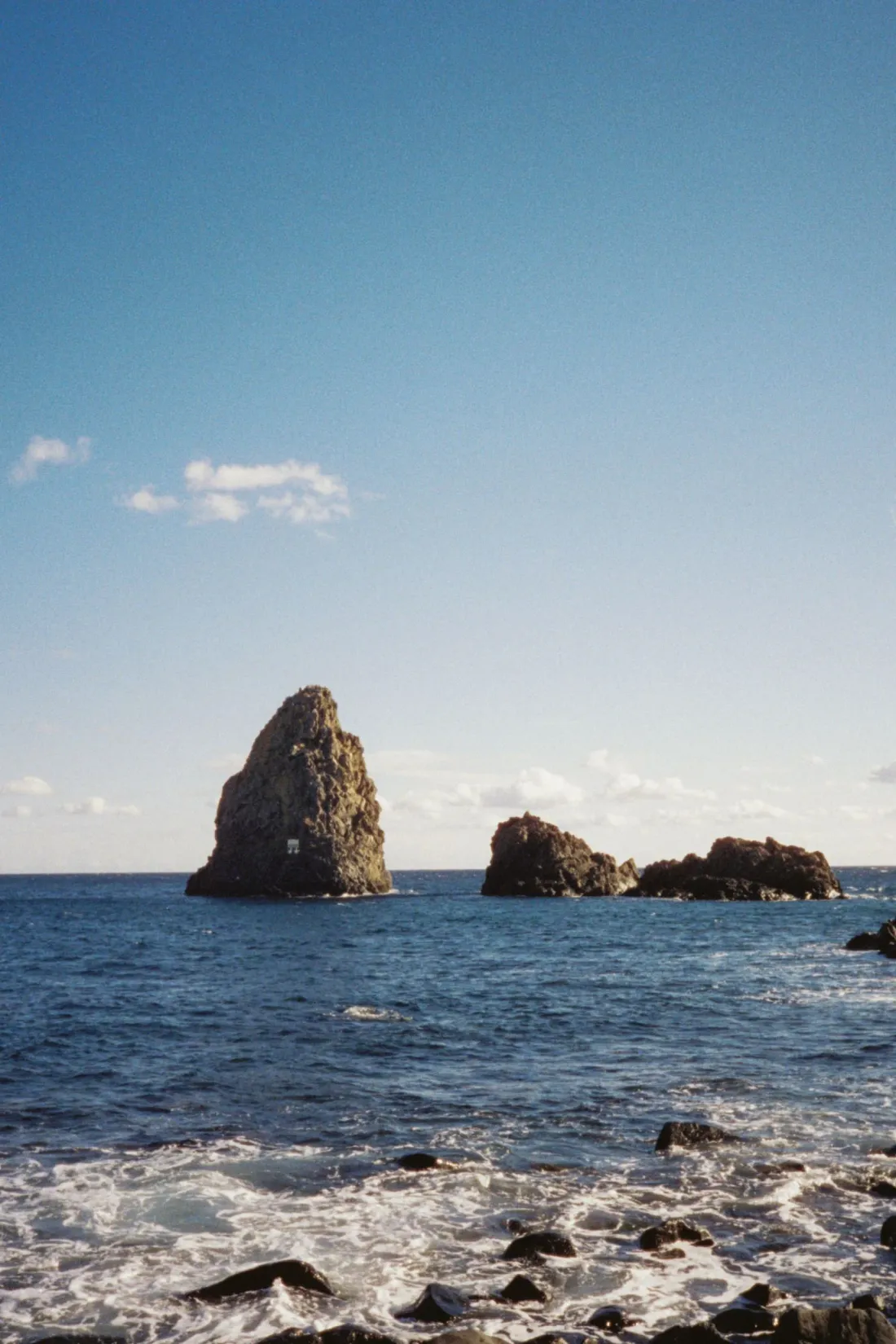 The basalt sea stacks of the Faraglioni dei Ciclopi rising from the blue water off Aci Trezza, Sicily, under a clear sky.
