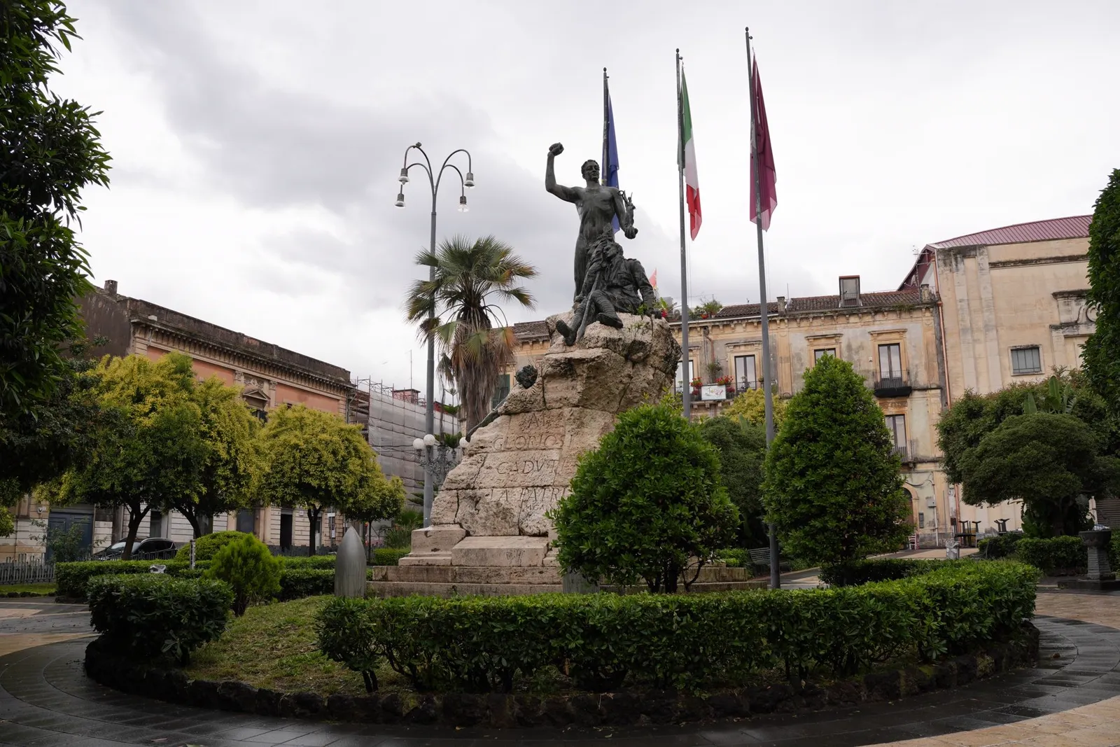 A bronze war memorial statue surrounded by palm trees and Baroque buildings in Piazza Garibaldi, Acireale, Sicily, on an overcast day.