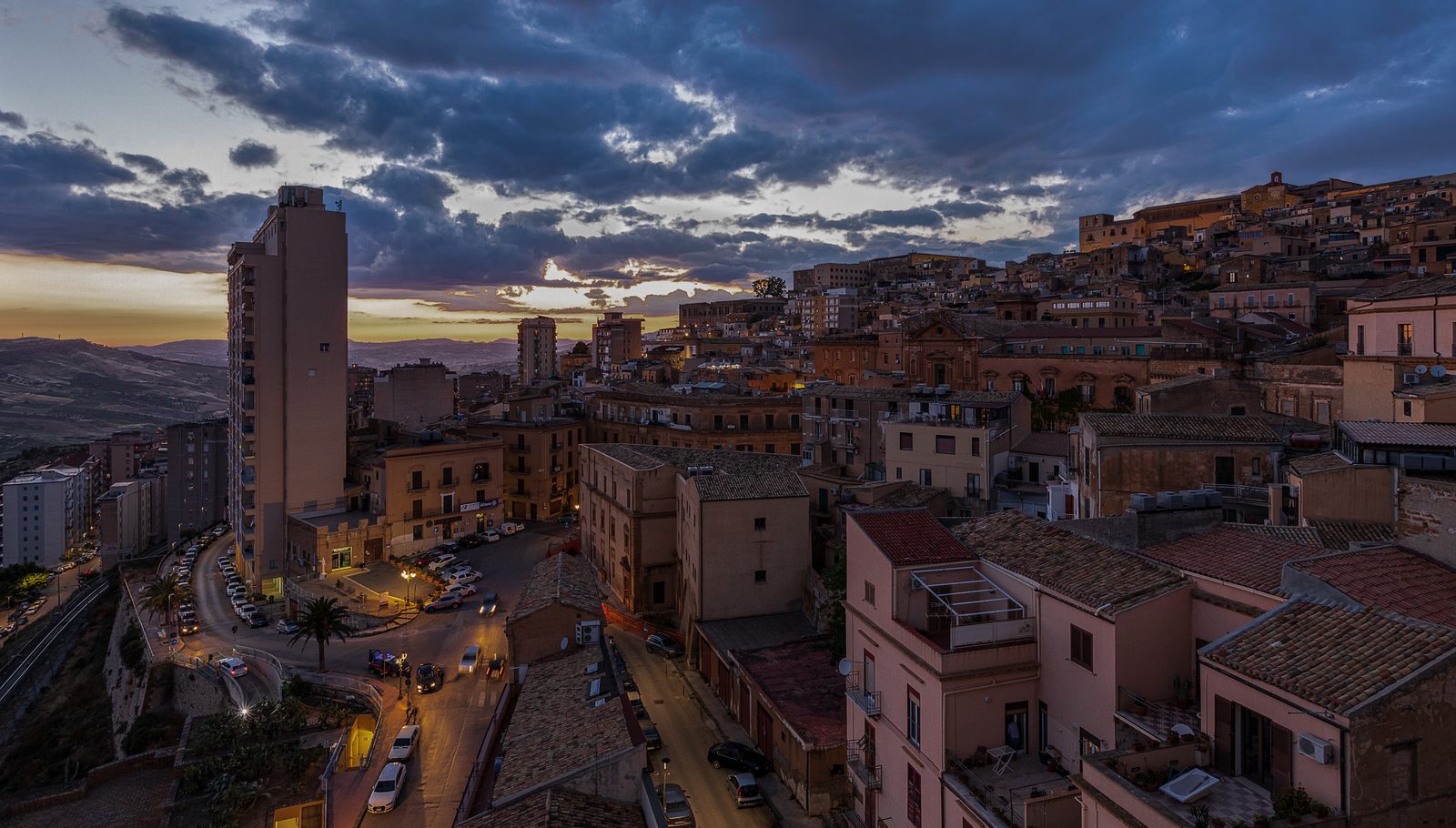 An elevated sunset view over the hillside old town of Agrigento, Sicily, with terracotta rooftops, the cathedral visible in the distance, and warm evening light over the surrounding hills.