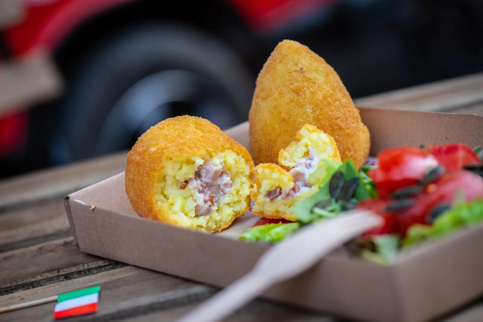 Two Sicilian arancini in a takeaway box, one broken open to show the rice, ragù and peas inside, with salad alongside