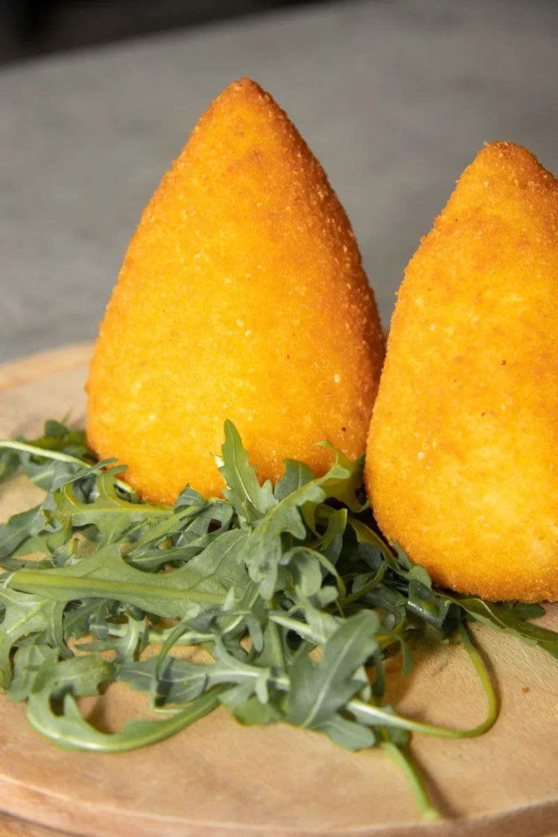 A cone-shaped arancino from Catania, fried golden brown, next to a second one on a wooden board with arugula.