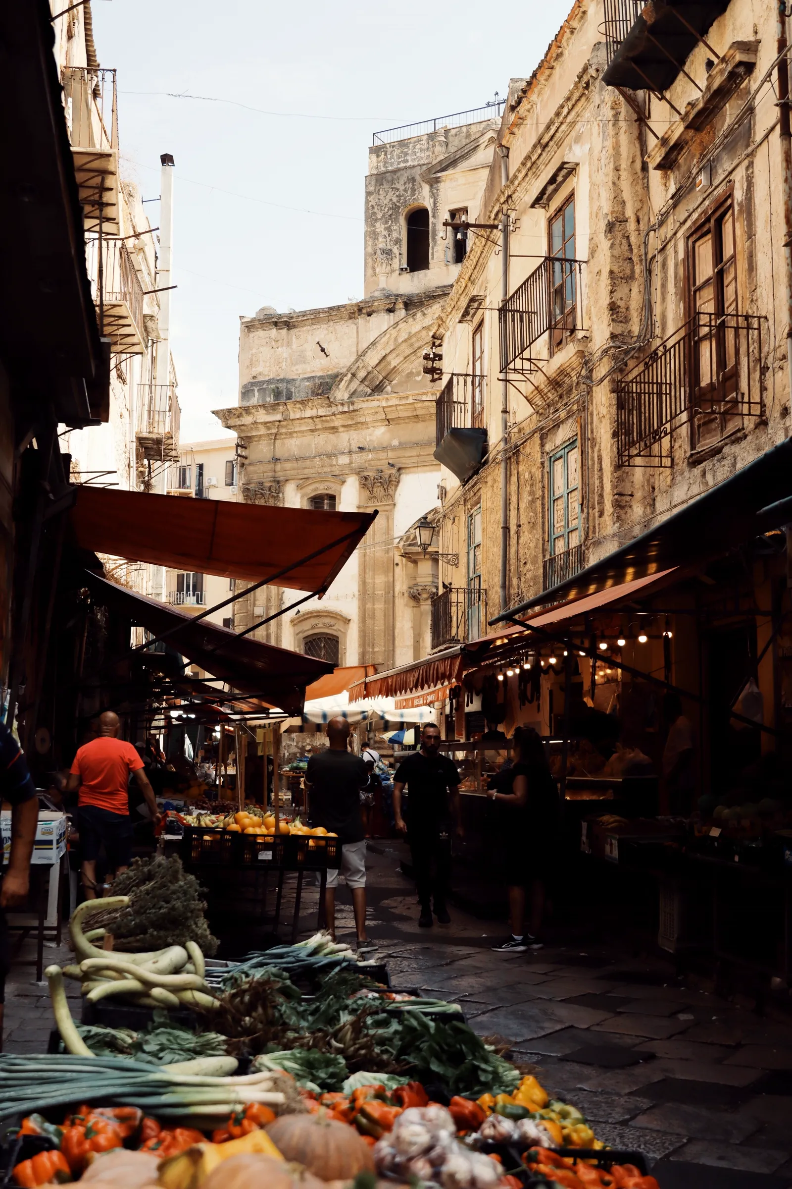A narrow Palermo market street lined with vegetable stalls piled high with fresh produce, awnings overhead and historic buildings on either side.