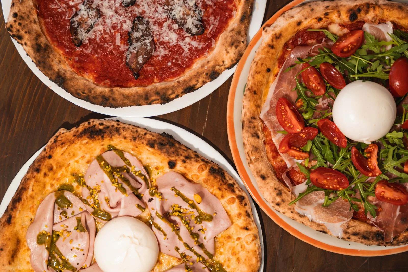 Three freshly baked pizzas with burrata, prosciutto, arugula, and mushrooms on a wooden table.