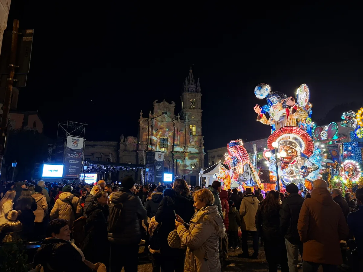 An illuminated, mechanically animated carnival float with giant papier-mâché figures in Piazza Duomo, Acireale, at night.