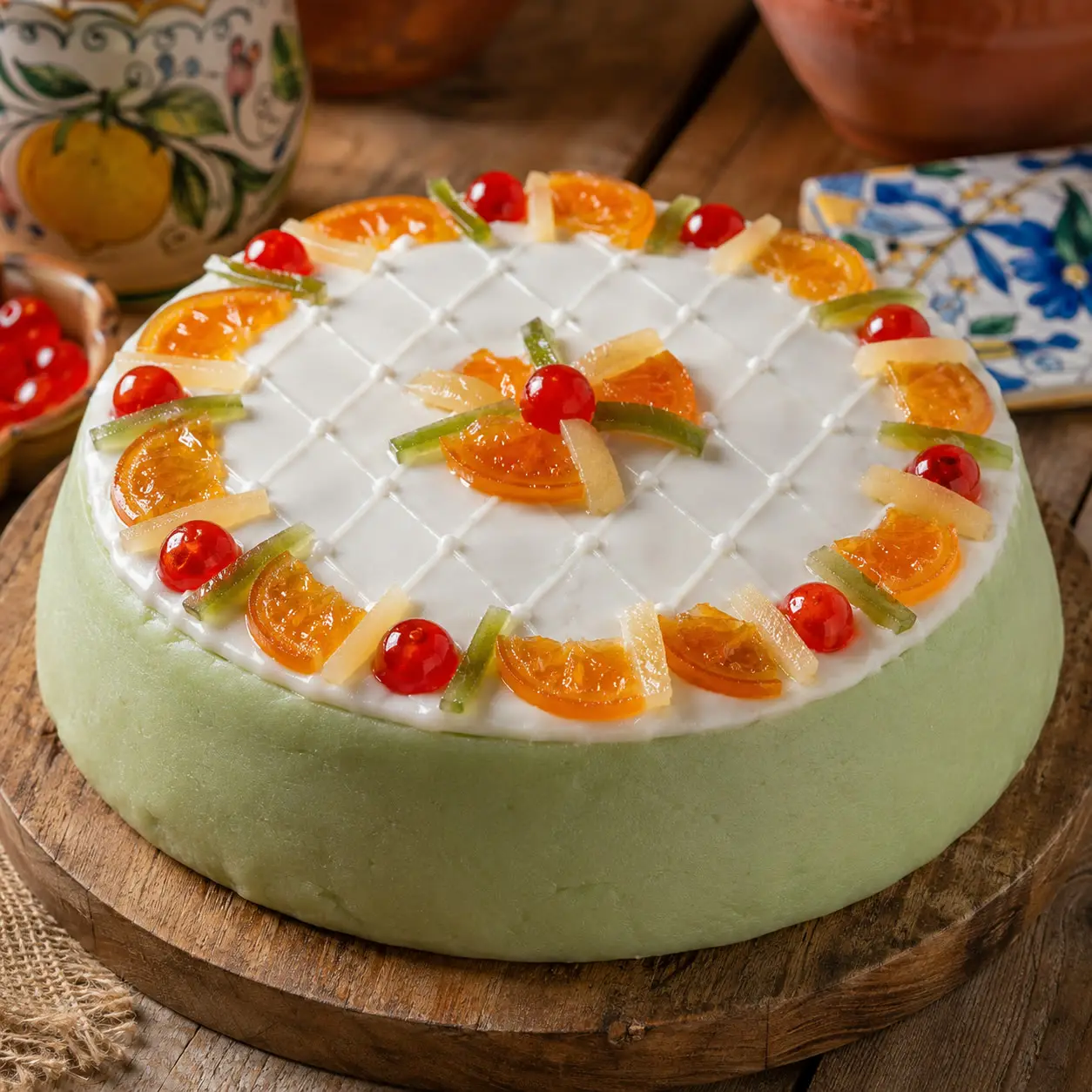 A whole Sicilian Cassata cake covered in pale green marzipan, topped with a white lattice-patterned icing and a ring of candied orange slices, cherries, and citron strips, on a rustic wooden board.