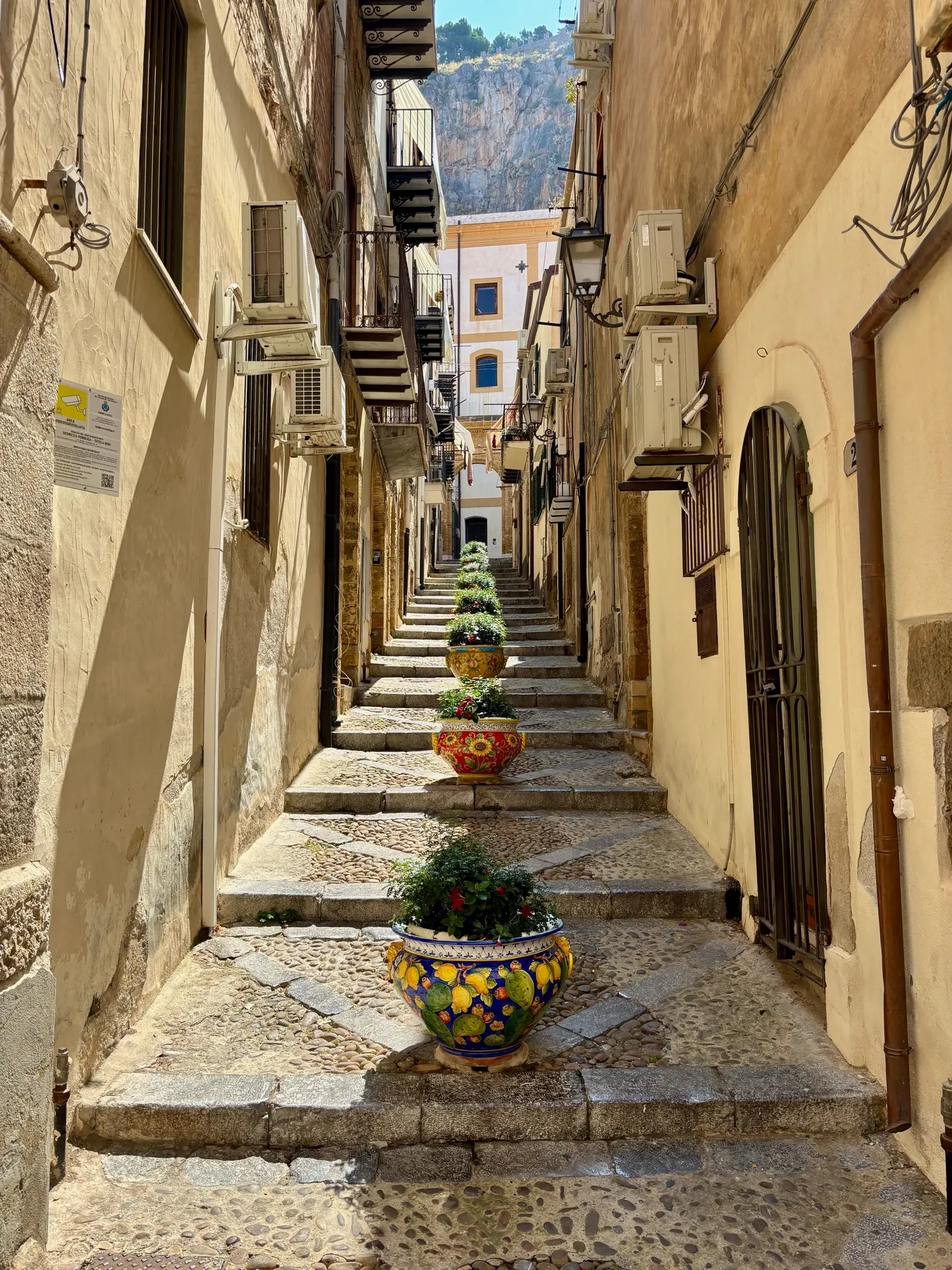 A stepped alley in Castelbuono's old town, lined with hand-painted ceramic pots and stone stairs leading up toward the Madonie mountains.