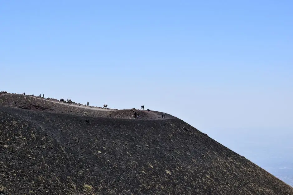 Hikers walking along the black volcanic crater rim of Mount Etna, Sicily, under a clear blue summer sky.