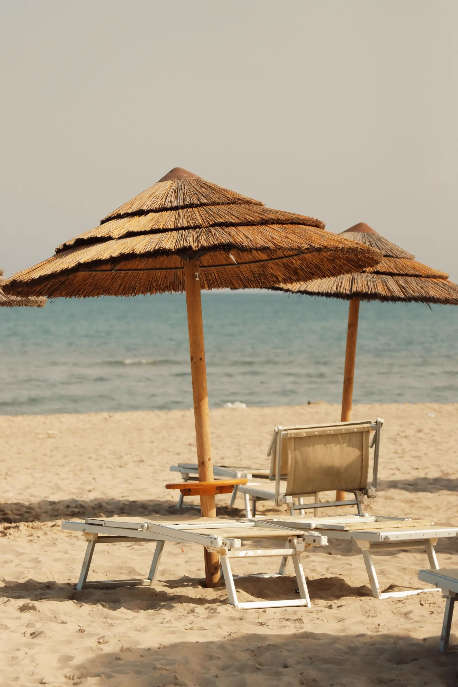 Traditional straw beach umbrellas and sun loungers on a golden-sand beach in Catania, Sicily, with the calm sea visible behind them.