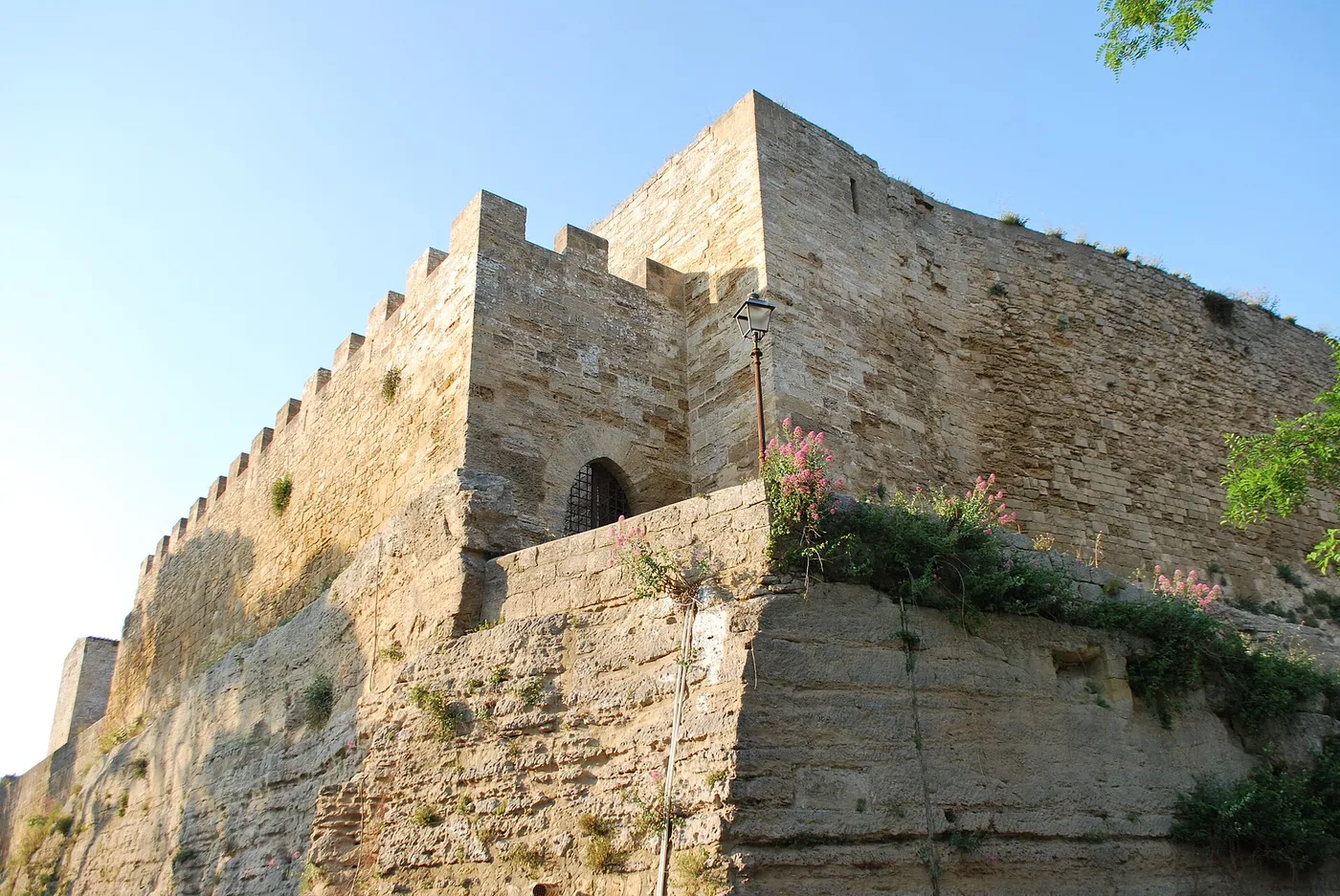 The crenellated stone walls of the Castello di Lombardia in Enna seen from below, rising directly out of the rock with wildflowers growing along the base