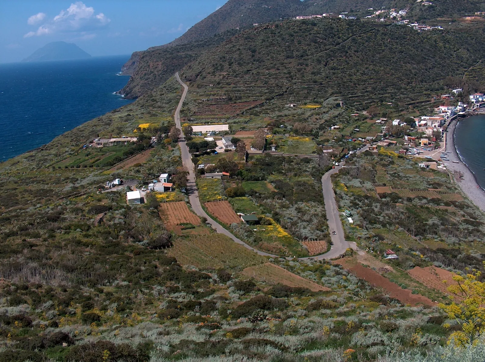 Terraced hillside on Filicudi dropping to the sea, a narrow road winding between scattered white houses and cultivated plots, with another Aeolian island on the horizon.