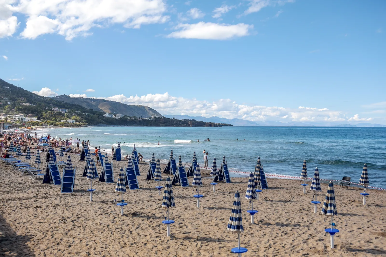 A long gold-sand beach in Giardini Naxos, Sicily, lined with rows of striped lido umbrellas and sunbeds, with a headland curving into the sea in the distance.