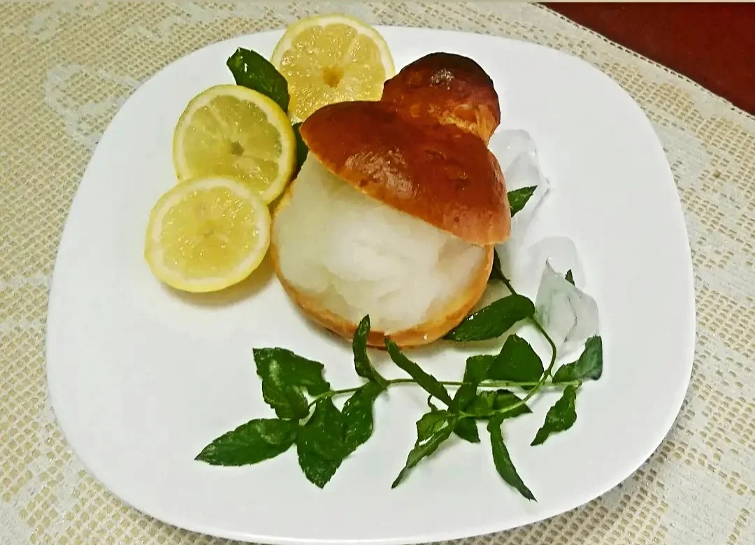 Lemon granita served inside a split brioche col tuppo, on a white plate with lemon slices and mint.