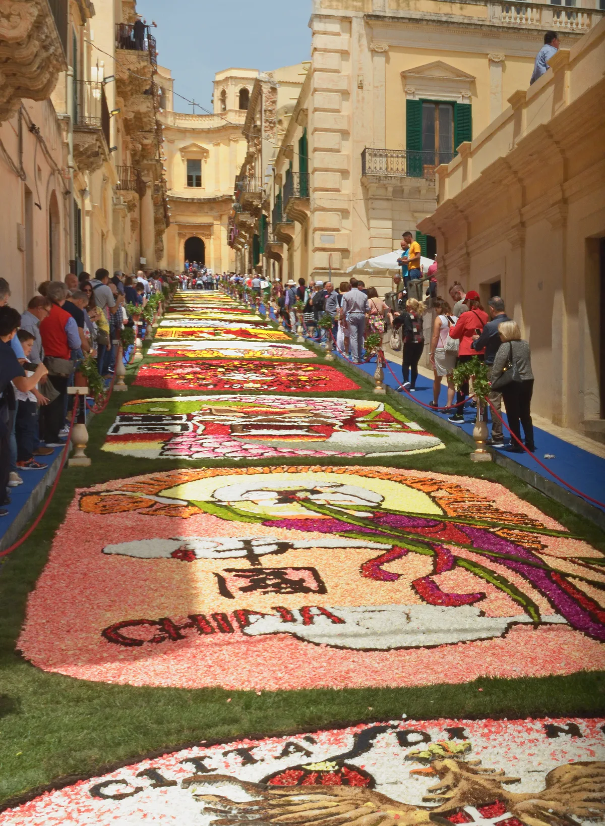 A long carpet of flower petals with intricate pictorial designs covering Via Nicolaci in Noto, lined with spectators.