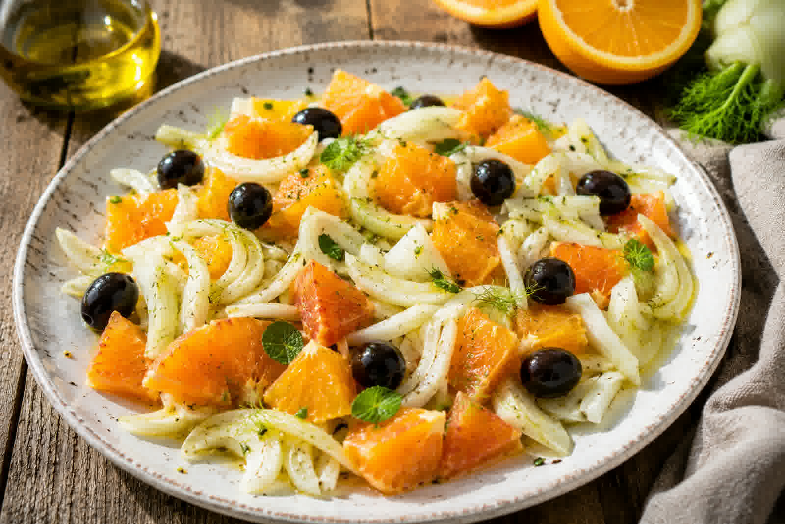 A Sicilian orange and fennel salad on a rustic plate, with thin slices of blood orange and raw fennel, black olives, and a drizzle of olive oil, photographed from above.