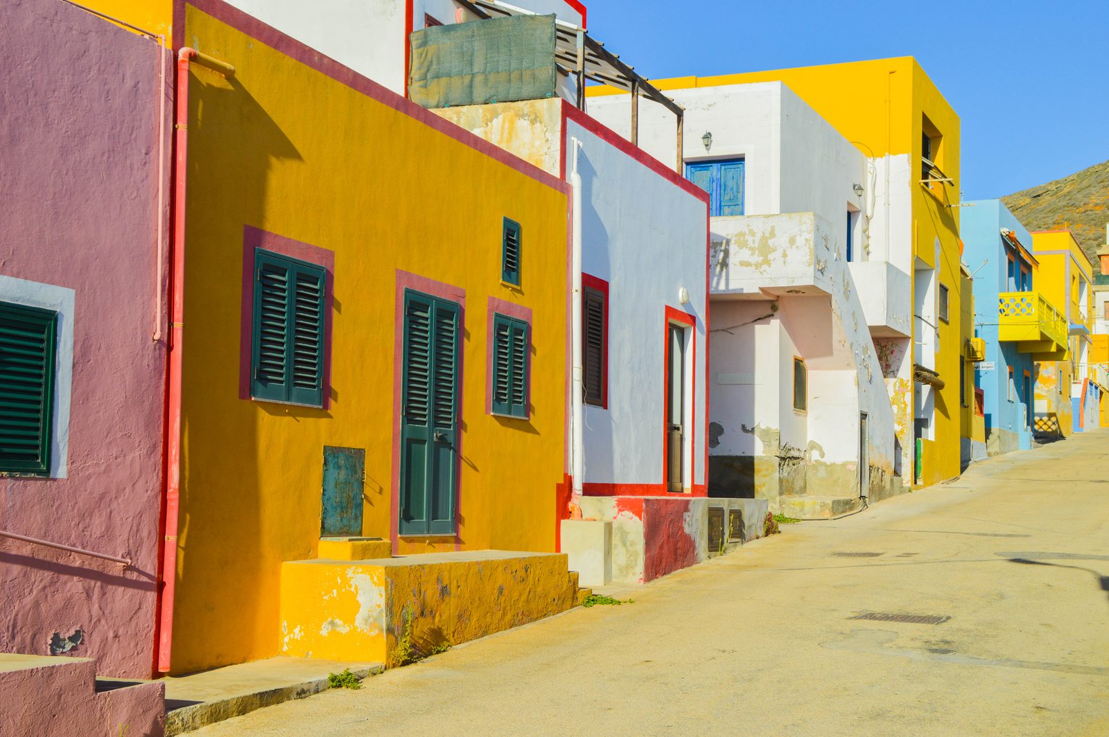 A street in Linosa's village lined with cube-shaped houses painted in bright yellow, pink, blue, and white, under a clear blue sky.