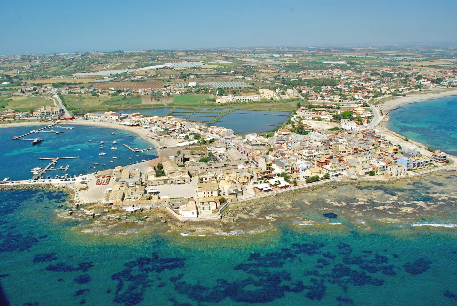 Aerial view of Marzamemi in Sicily, the low village on its small peninsula surrounded by turquoise shallows, with fishing boats moored along the shore.