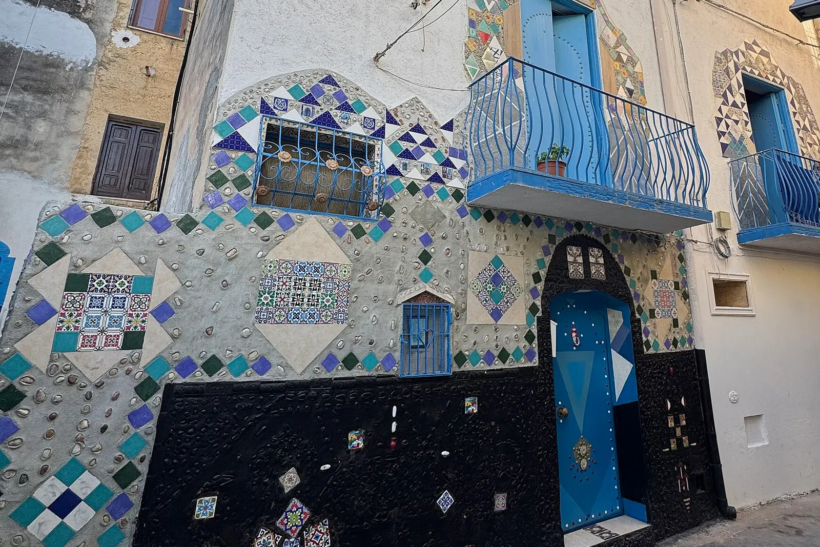 A house front in the Kasbah district of Mazara del Vallo, Sicily, covered in hand-set ceramic mosaic — coloured diamonds, patterned maiolica tiles and shells — with a Moorish-arched blue door and blue wrought-iron balconies above.