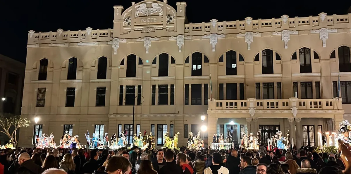 A nighttime procession of ornate sacred sculptural groups in front of an illuminated historic building in Trapani.