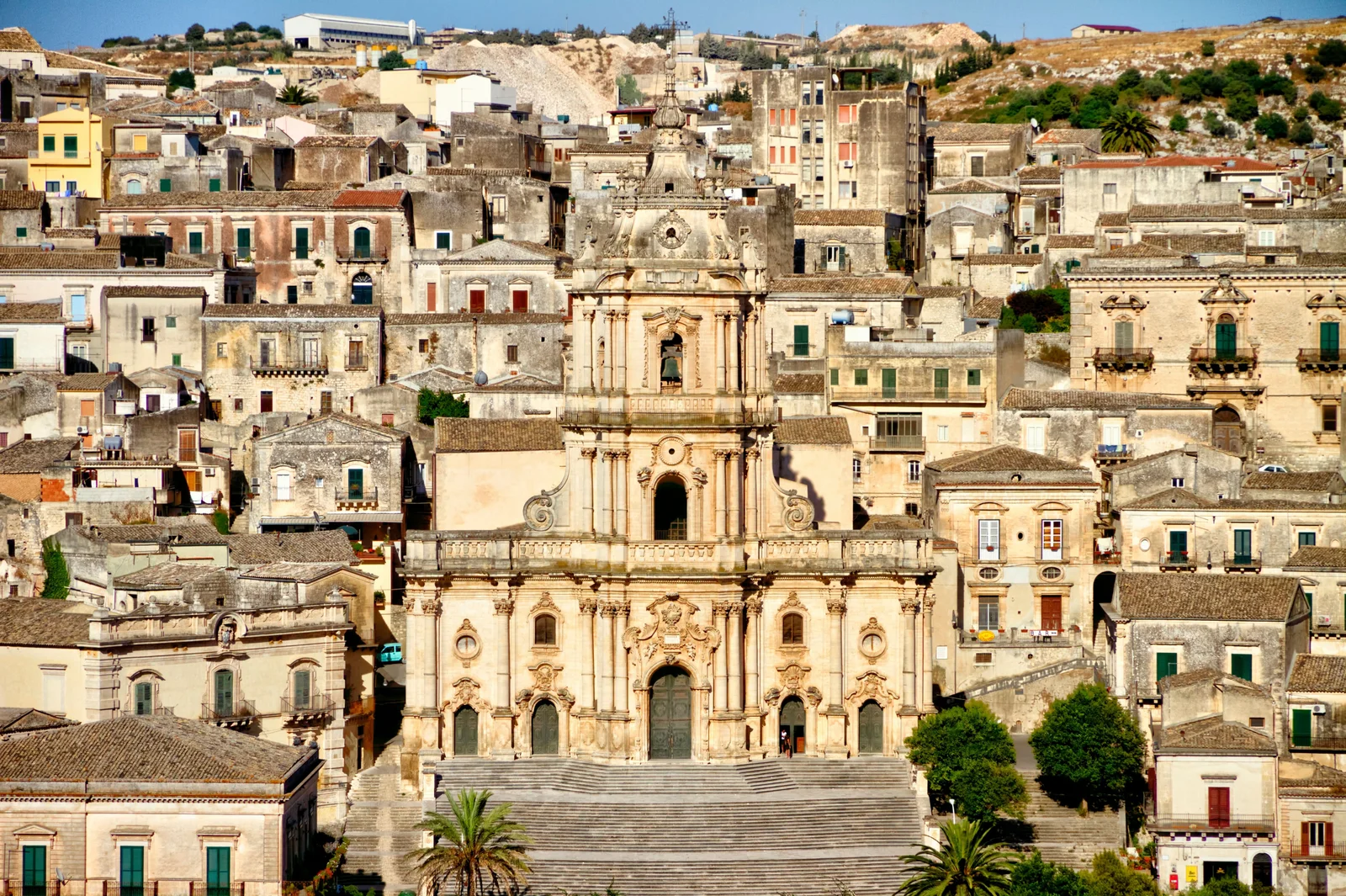 Baroque facade of the Duomo di San Giorgio in Modica with a grand staircase, surrounded by the dense hillside old town of golden stone.