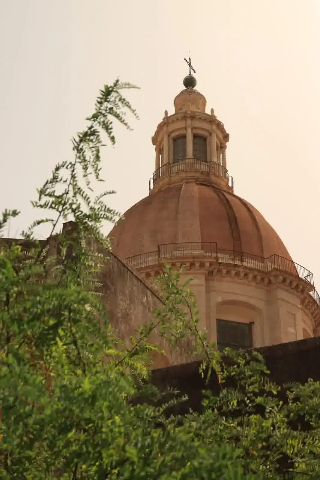 The large Baroque dome and lantern of the Monastero dei Benedettini church of San Nicolò l'Arena in Catania, Sicily, seen from below through green foliage.
