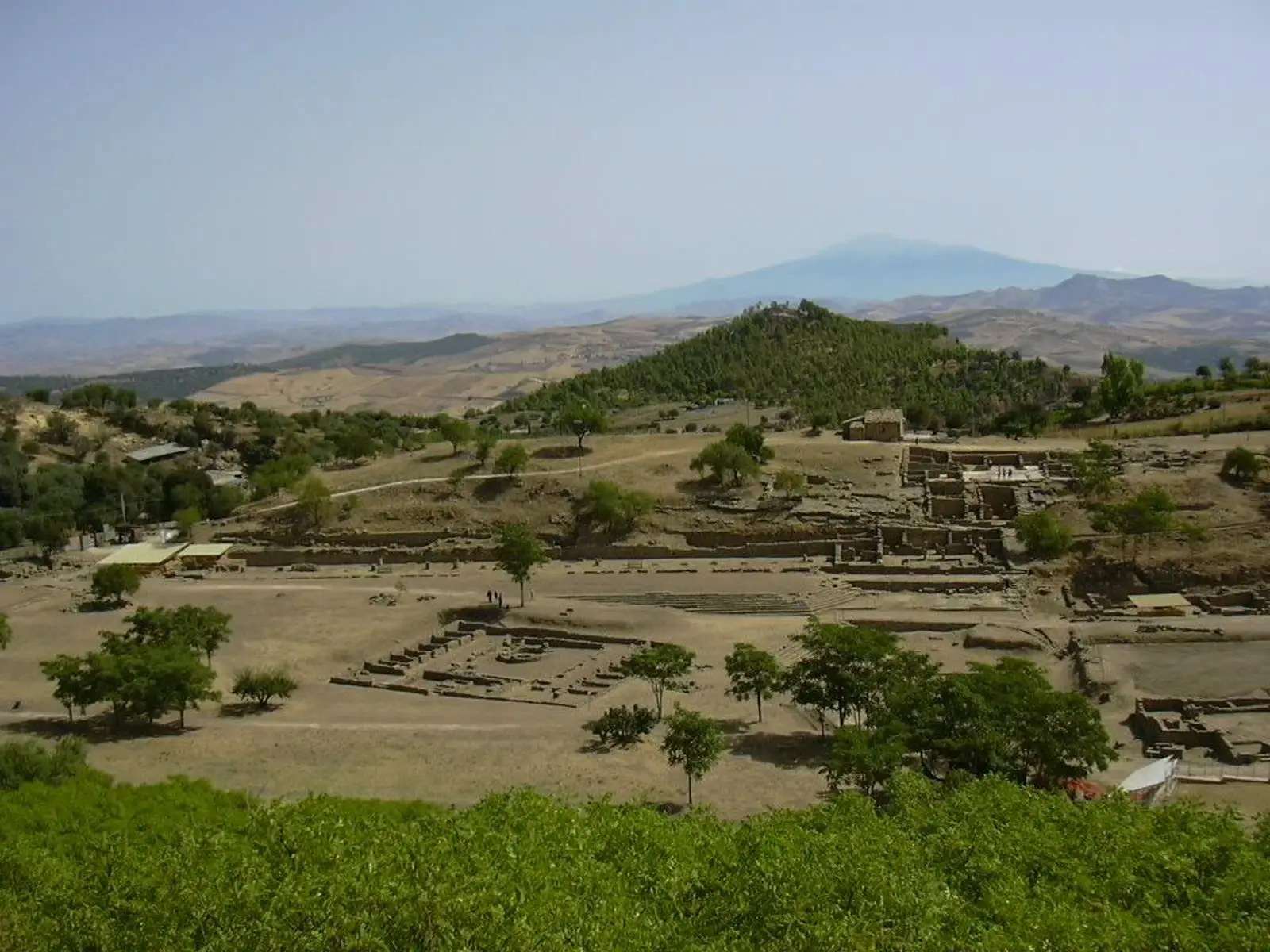 A wide view over the excavated ruins of Morgantina's agora and theater from a hillside, with Mount Etna visible on the horizon.