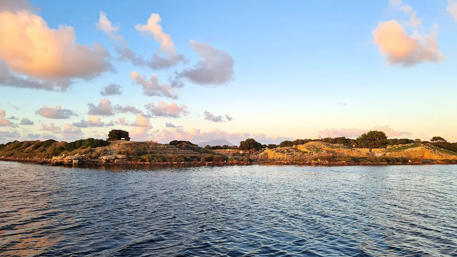 The low island of Mozia (San Pantaleo) seen from the water at golden hour in the Stagnone lagoon near Marsala, Sicily, with ancient stone walls and excavated ruins visible along the shoreline under a pale evening sky.