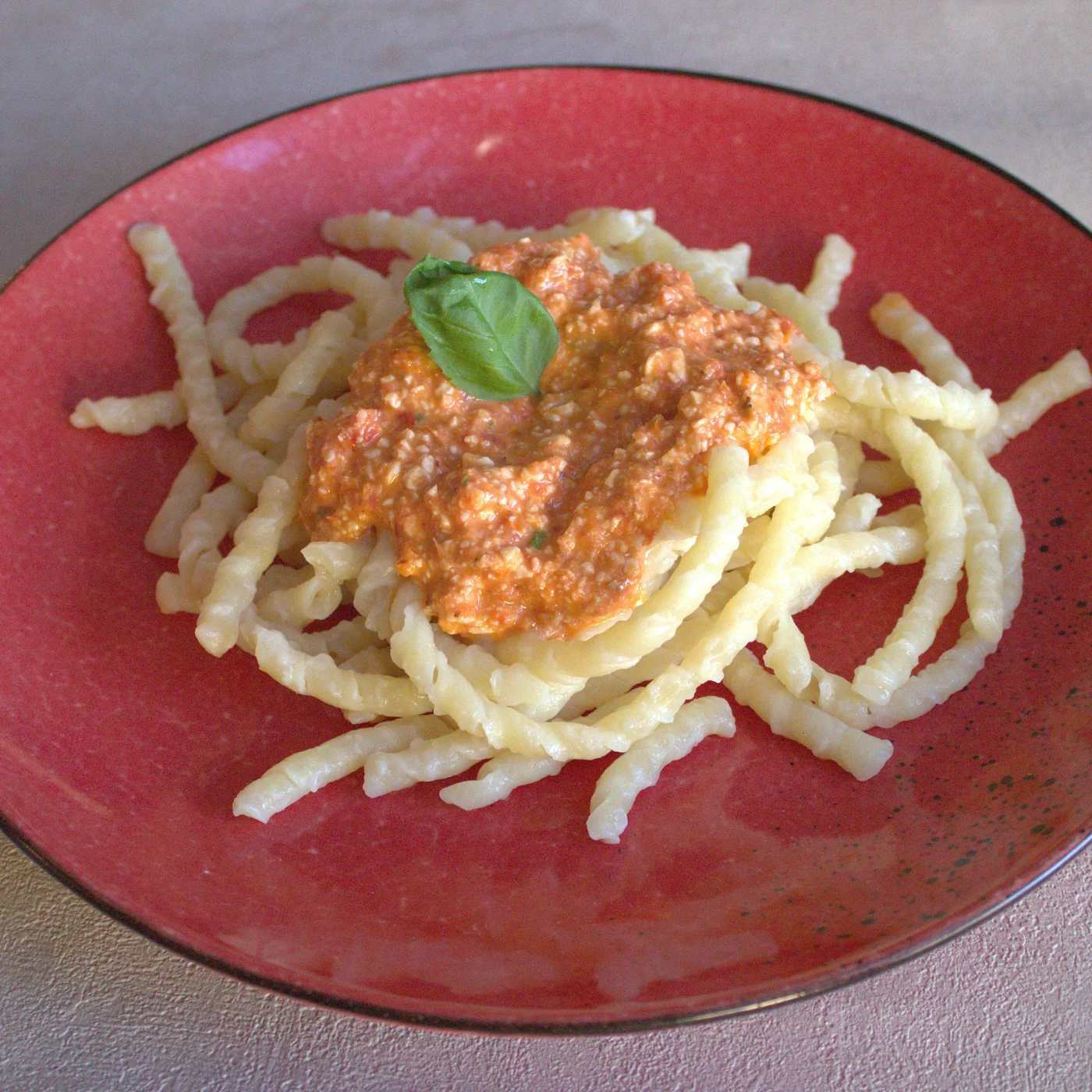 Busiate with Pesto alla Trapanese on a red plate, garnished with a basil leaf — the coarse, reddish tomato-and-almond sauce sits visibly chunky on top of the pasta.