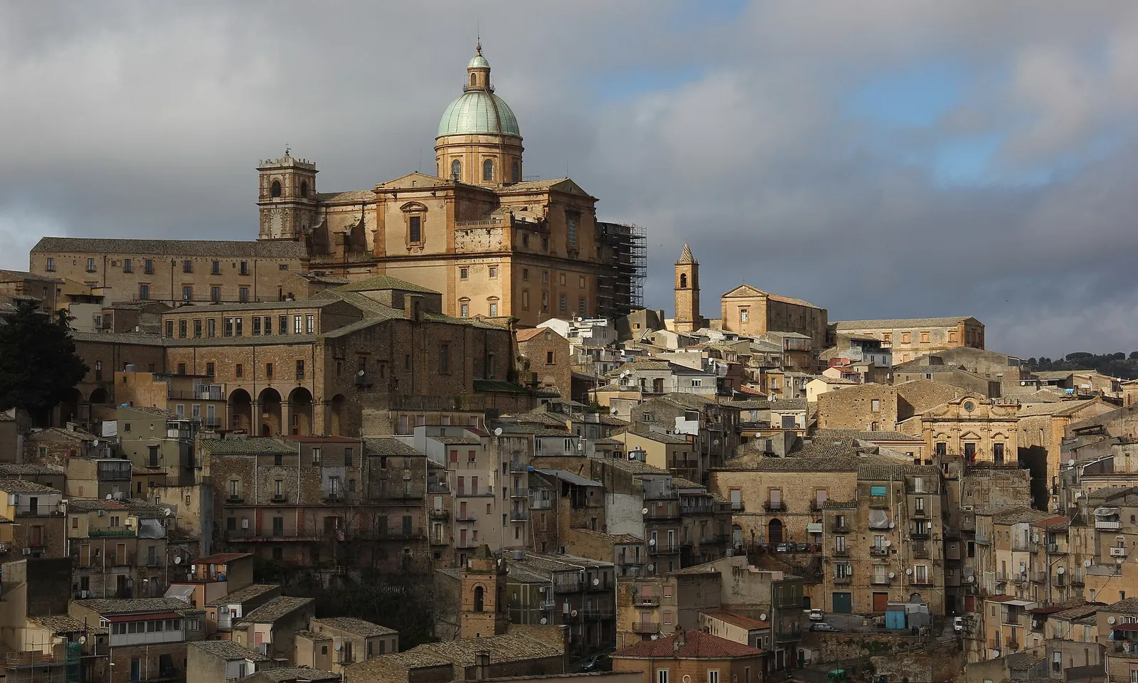 The cathedral of Piazza Armerina in Sicily rising above the tightly stacked ochre houses of the old town on its hillside, its green dome catching the light under a heavy sky.