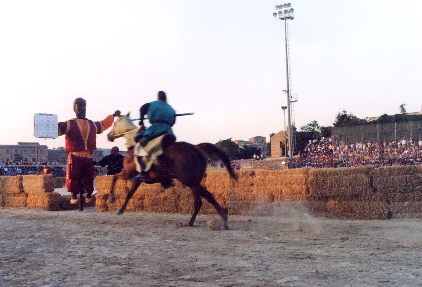 A rider in blue medieval costume charging on horseback at the Quintana target during the Palio dei Normanni in Piazza Armerina, with straw bales and a crowd behind