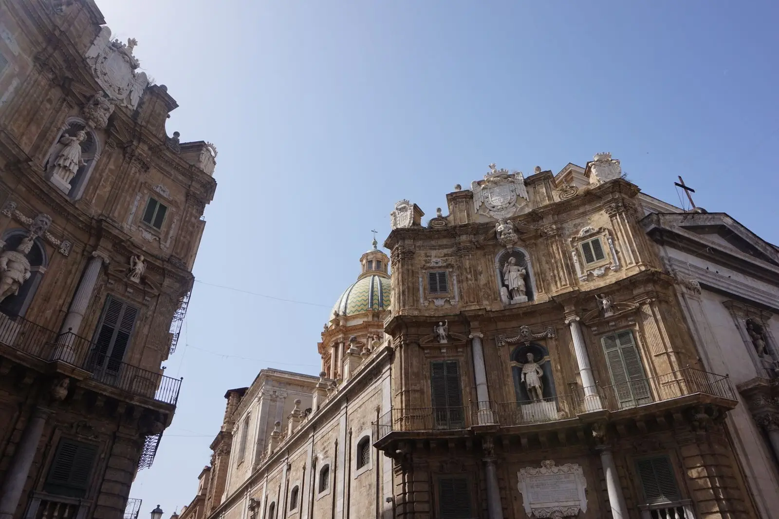 Two curved Baroque facades of Quattro Canti in Palermo meeting at a street corner, decorated with statues in niches and a green dome visible behind, under a clear blue sky.