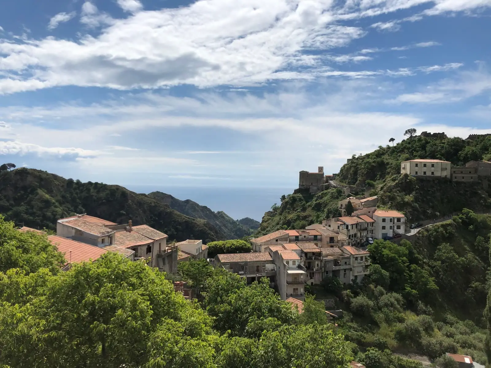 A hilltop Sicilian village with terracotta rooftops spilling down a green ridge, a stone castle tower perched on the crest above it, and the sea visible in the distance.