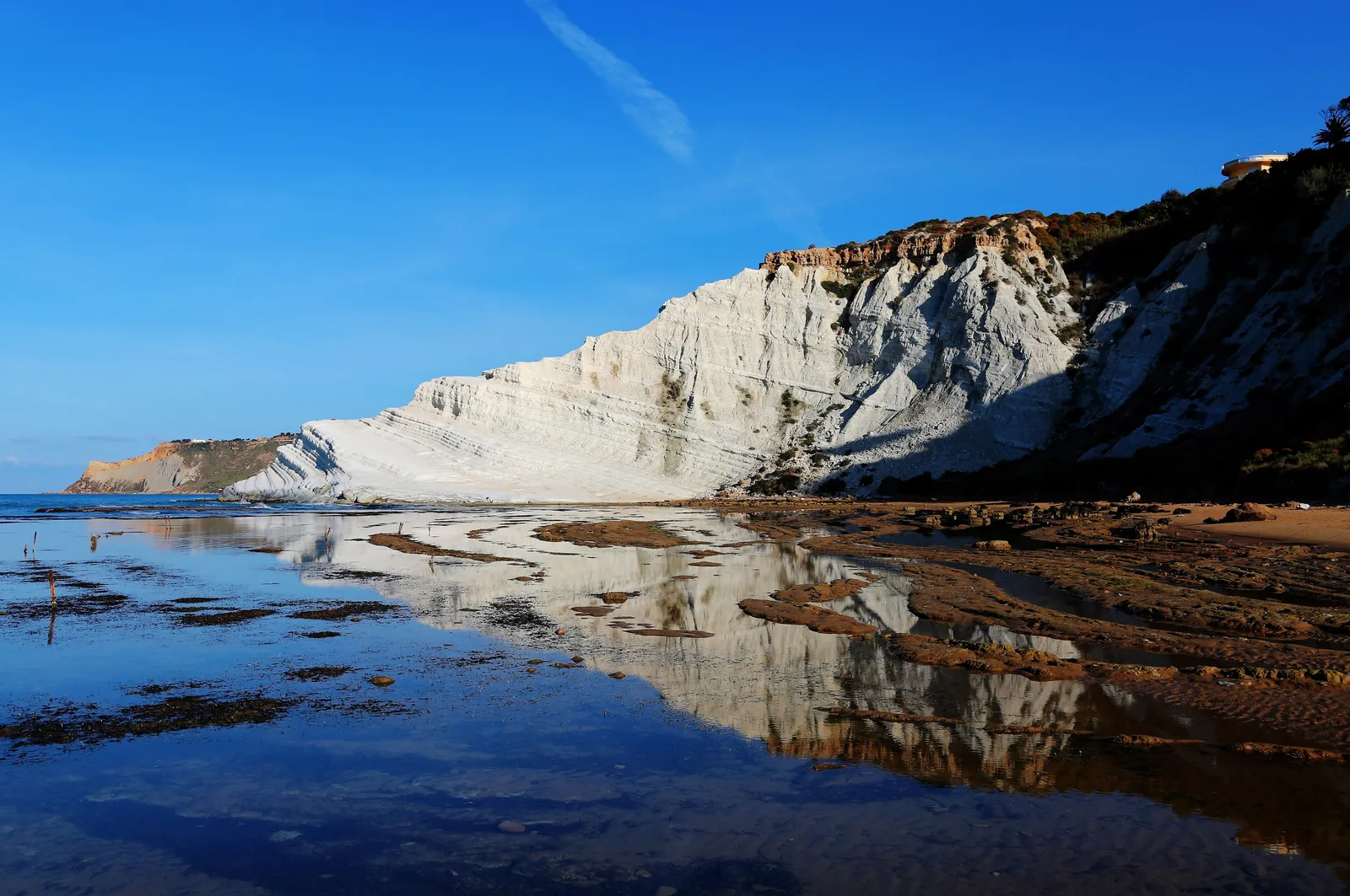 The white marl cliff of Scala dei Turchi near Realmonte, Sicily, its stepped terraces glowing in low sunlight and mirrored in a shallow pool of water on the rocky foreshore, under a deep blue sky.