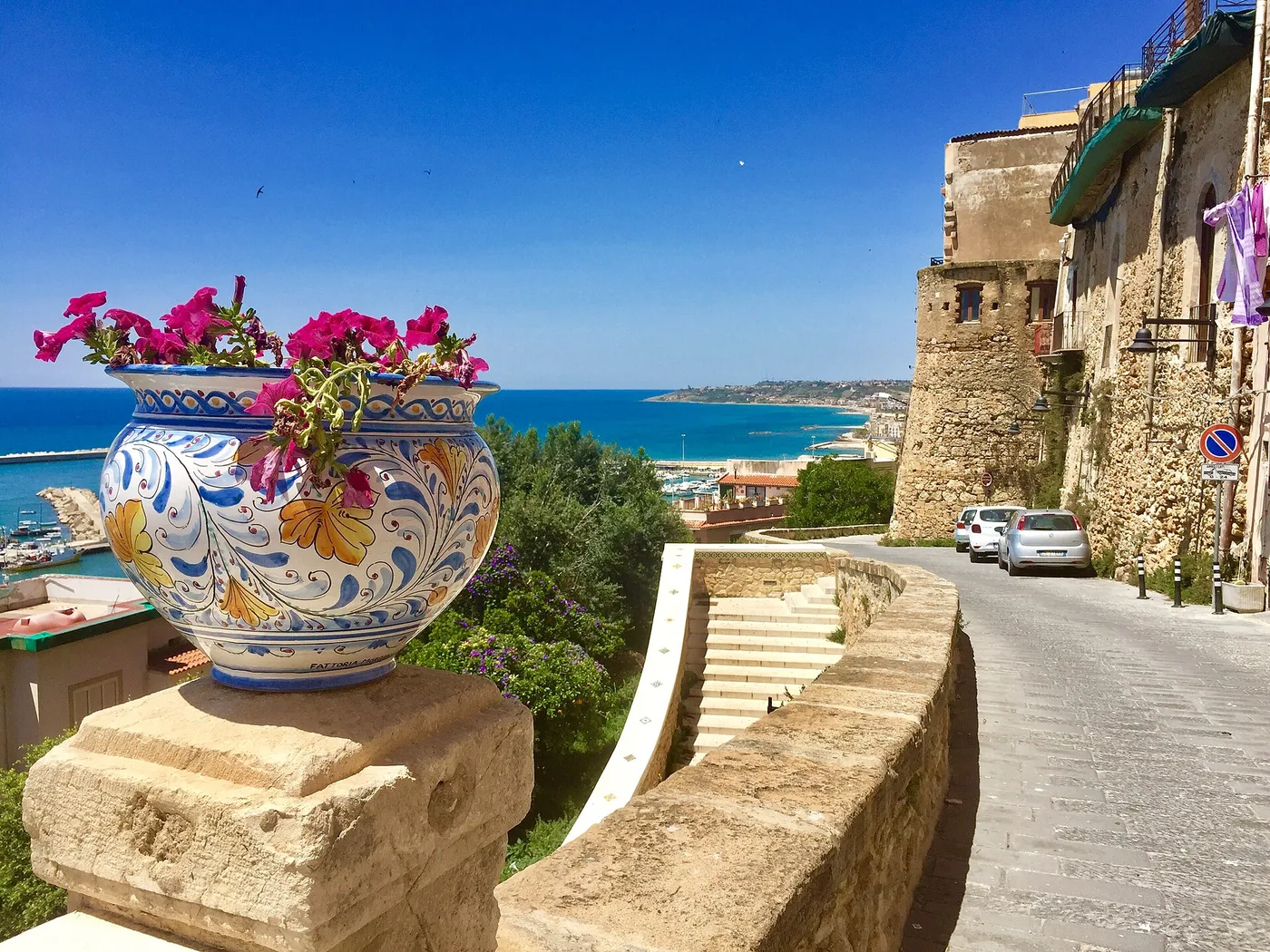 A large hand-painted Sciacca ceramic vase in yellow, blue and green, planted with pink flowers, standing on a stone plinth above the sea