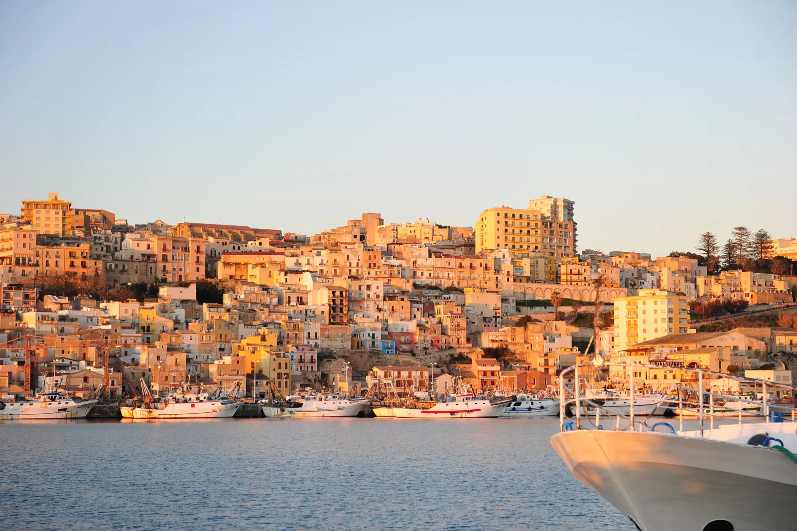 The town of Sciacca in Sicily seen from the water at golden hour, its honey-coloured houses stacked up the hillside above the harbour, with the fishing fleet moored along the quay in the foreground.