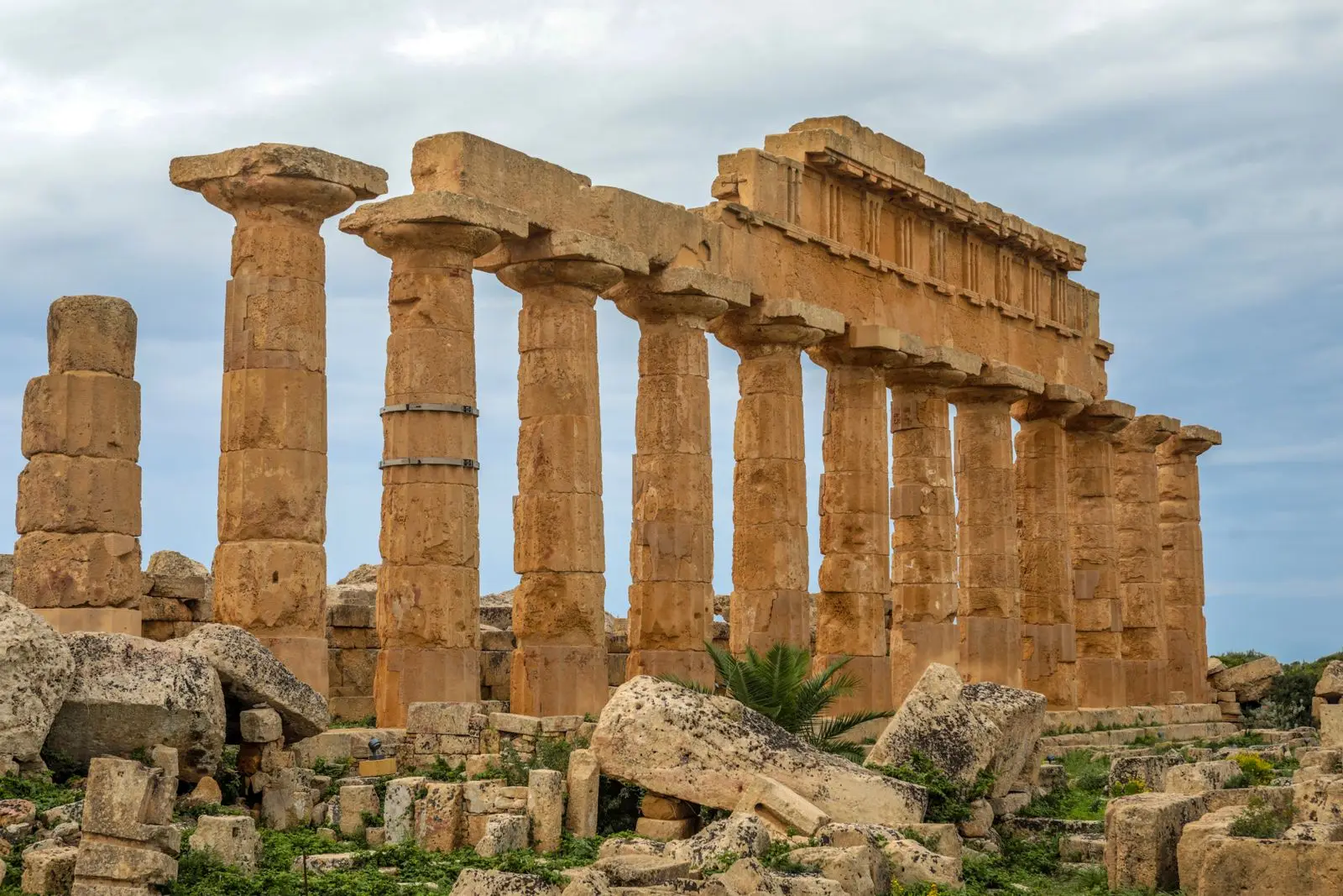 Weathered Doric columns and a partial entablature of Temple C on the acropolis of Selinunte, Sicily, standing among fallen stone blocks under a pale blue sky.