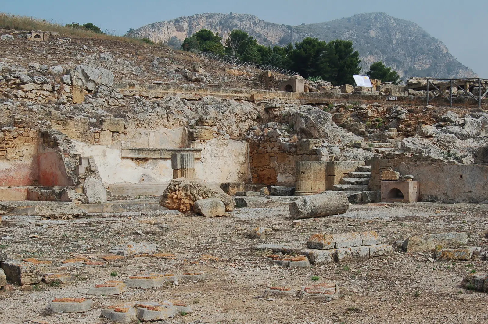 Excavated house remains at Solunto in Sicily, with stone walls, column bases and paved floors, and the slope of Monte Catalfano rising behind