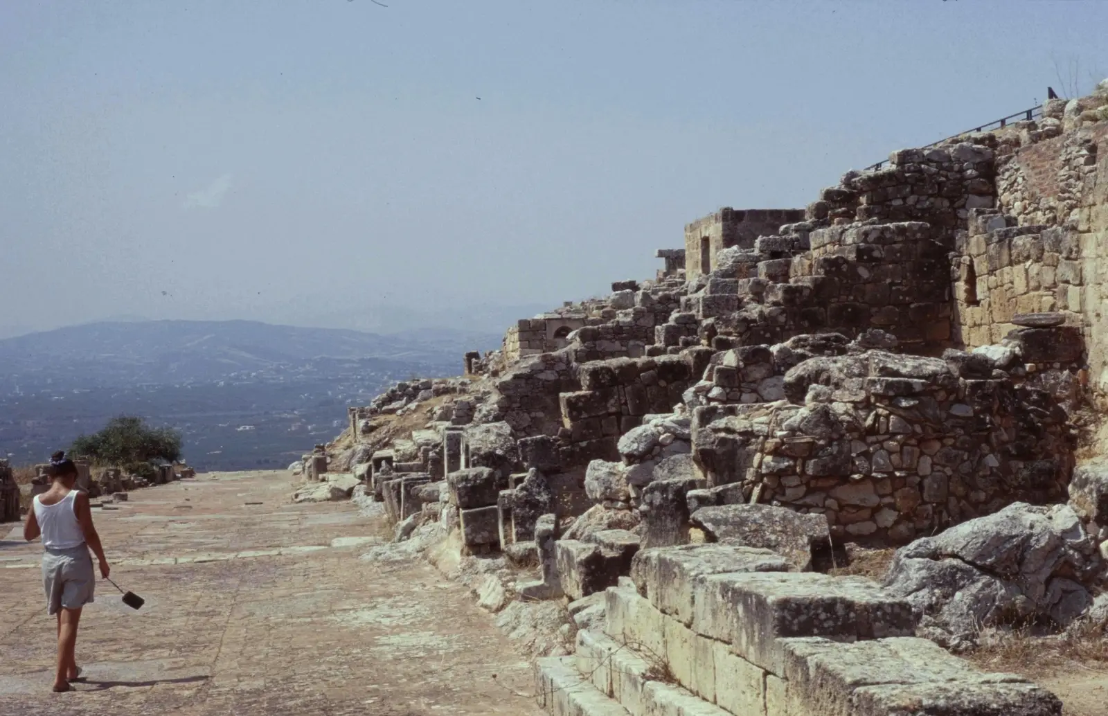 The paved main street of the ancient city of Solunto in Sicily, climbing between the ruined stone fronts of Hellenistic houses with the coastal plain hazy beyond.