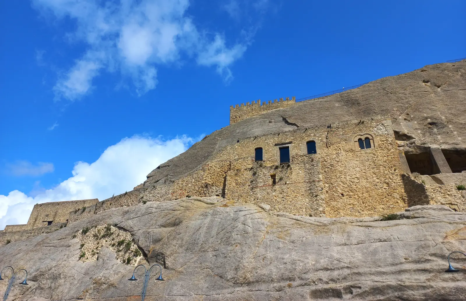 The rock castle of Sperlinga in Sicily, its windows and rooms cut directly into a pale sandstone outcrop beneath battlements, against a blue sky.