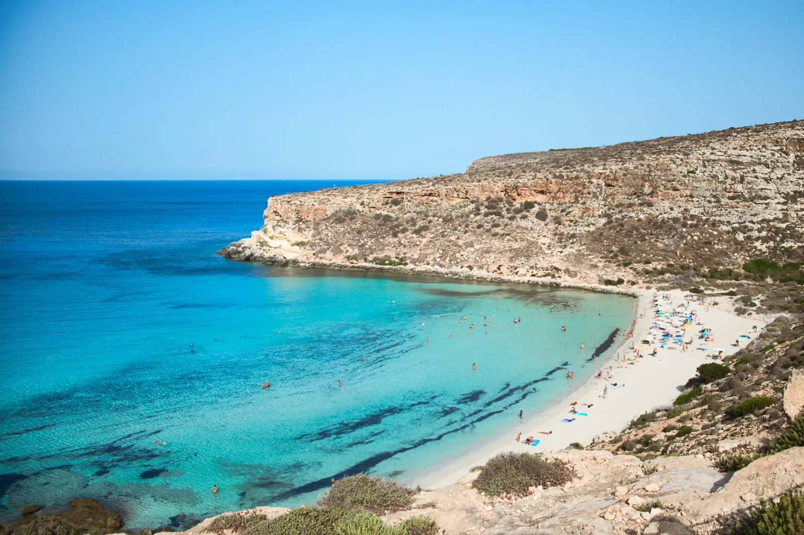 Aerial view of Spiaggia dei Conigli on Lampedusa, showing a crescent of white sand and exceptionally clear turquoise-to-deep-blue water surrounded by pale rocky cliffs.