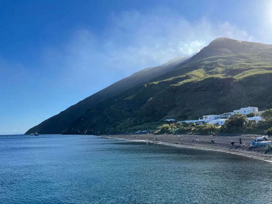 View from the sea toward Stromboli: a green volcanic slope drops steeply to the water, a small white village sits on the shore, with clear turquoise water in front.