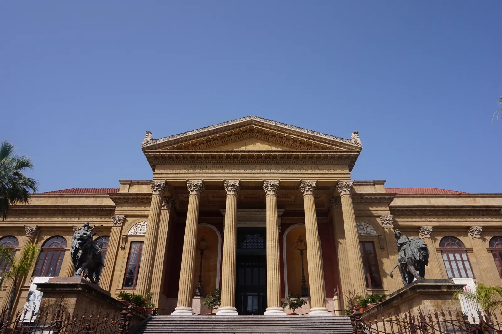The grand neoclassical facade of Teatro Massimo in Palermo, with its wide front steps, Corinthian columns and bronze lion statues under a clear blue sky.