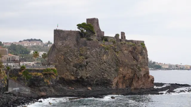 The black lava Norman castle of Aci Castello perched on a rocky basalt promontory over the sea near Catania, Sicily, with the coastline stretching behind it.