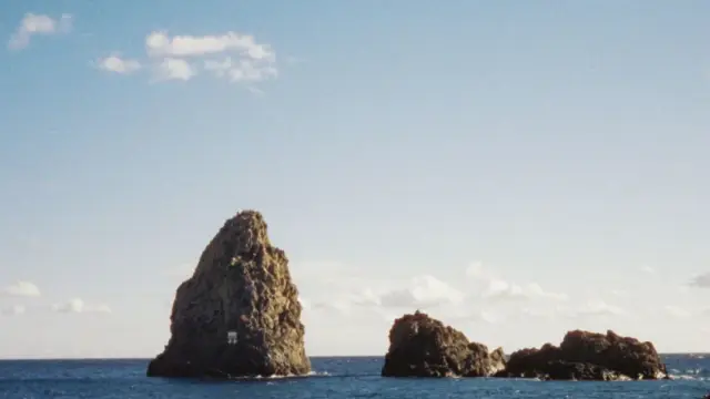 The basalt sea stacks of the Faraglioni dei Ciclopi rising from the blue water off Aci Trezza, Sicily, under a clear sky.