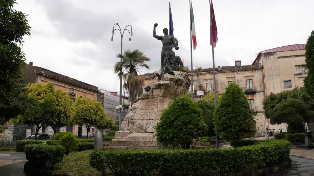 A bronze war memorial statue surrounded by palm trees and Baroque buildings in Piazza Garibaldi, Acireale, Sicily, on an overcast day.