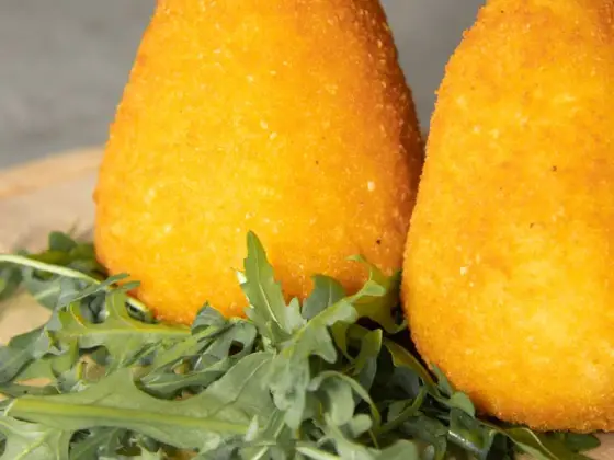 A cone-shaped arancino from Catania, fried golden brown, next to a second one on a wooden board with arugula.