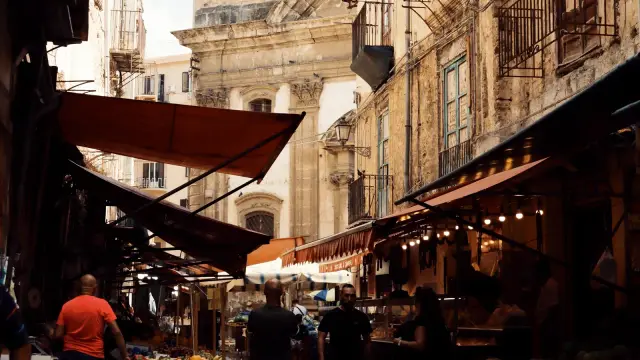 A narrow Palermo market street lined with vegetable stalls piled high with fresh produce, awnings overhead and historic buildings on either side.