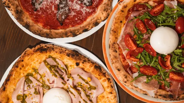 Three freshly baked pizzas with burrata, prosciutto, arugula, and mushrooms on a wooden table.