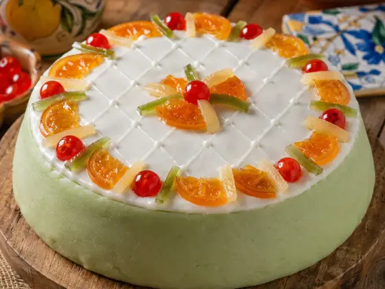 A whole Sicilian Cassata cake covered in pale green marzipan, topped with a white lattice-patterned icing and a ring of candied orange slices, cherries, and citron strips, on a rustic wooden board.