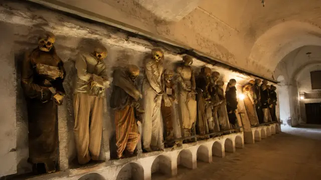 A row of mummified figures dressed in period clothing standing in wall niches along a vaulted stone corridor in the Capuchin Catacombs of Palermo.